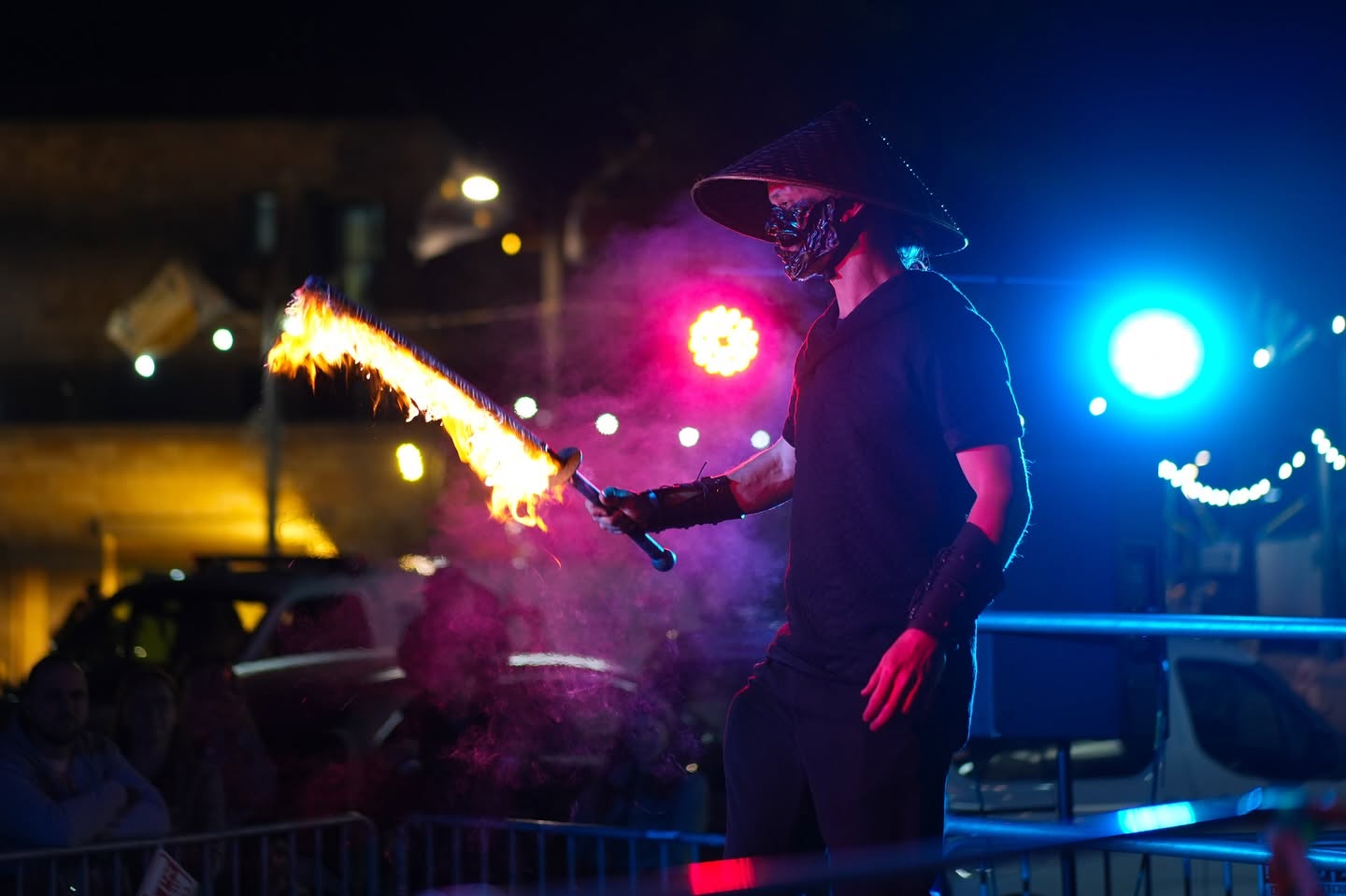 A performer breathes fire at night with coloured lights behind him.