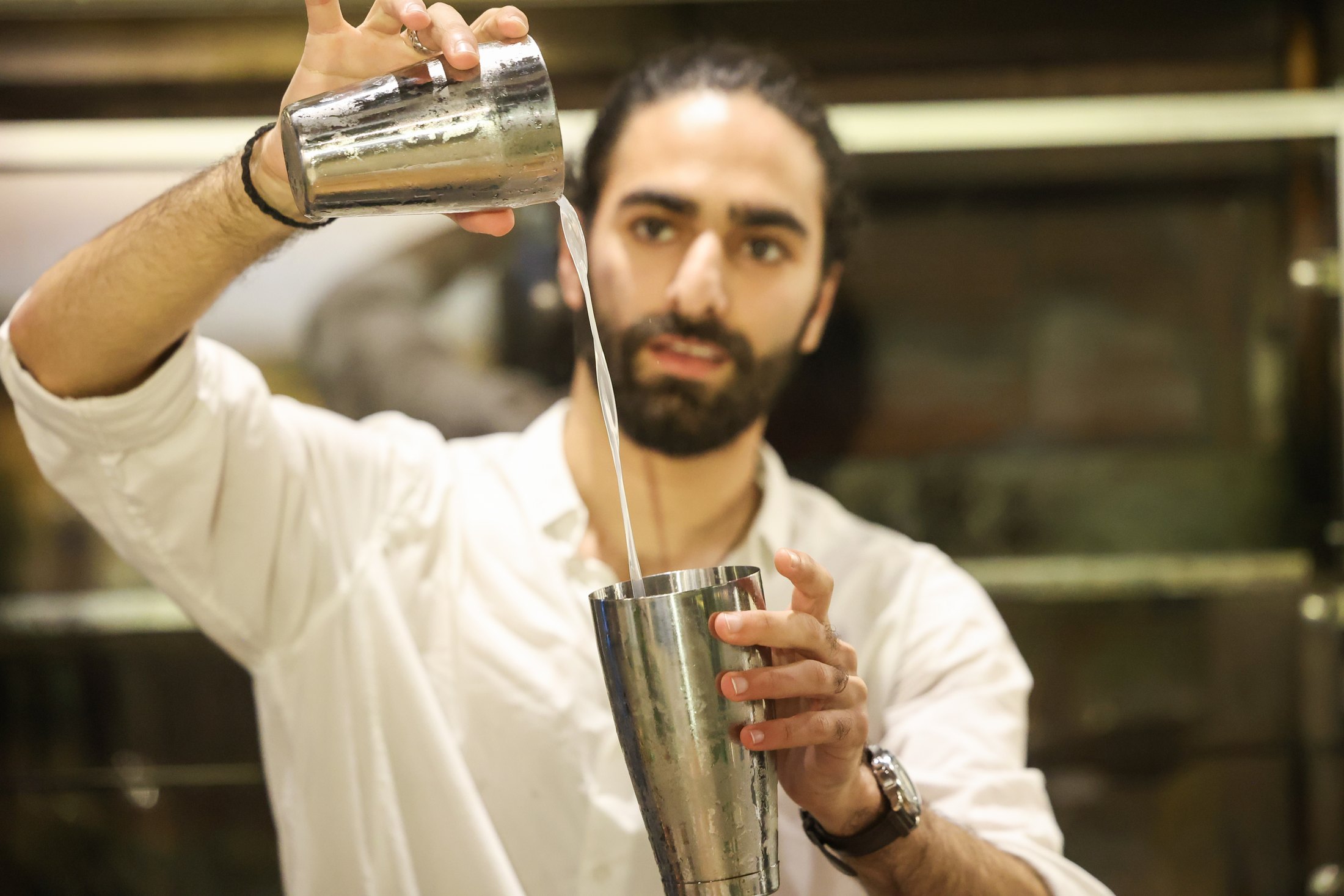 A bartender pours a drink between two mixing vessels.