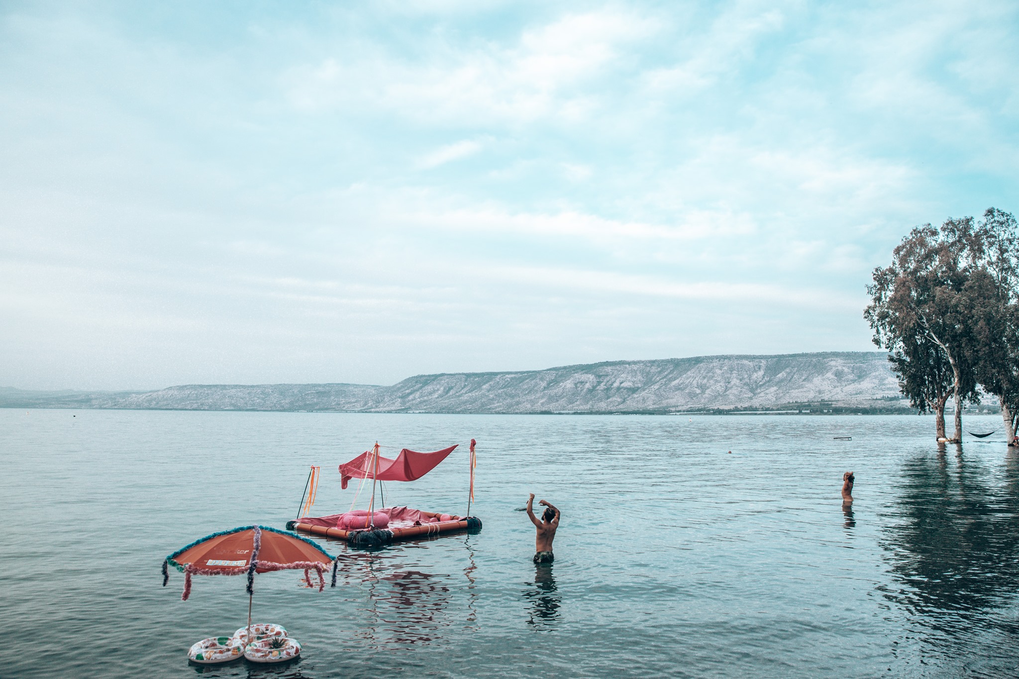 People swim in the Sea of Galilee near a pink raft and trees in the water.