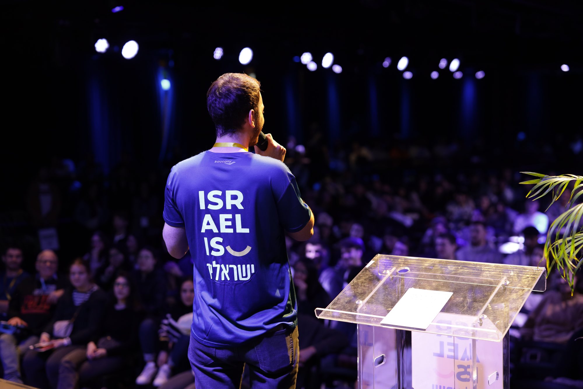 Rear view of a speaker in an Israel-is shirt facing the audience.