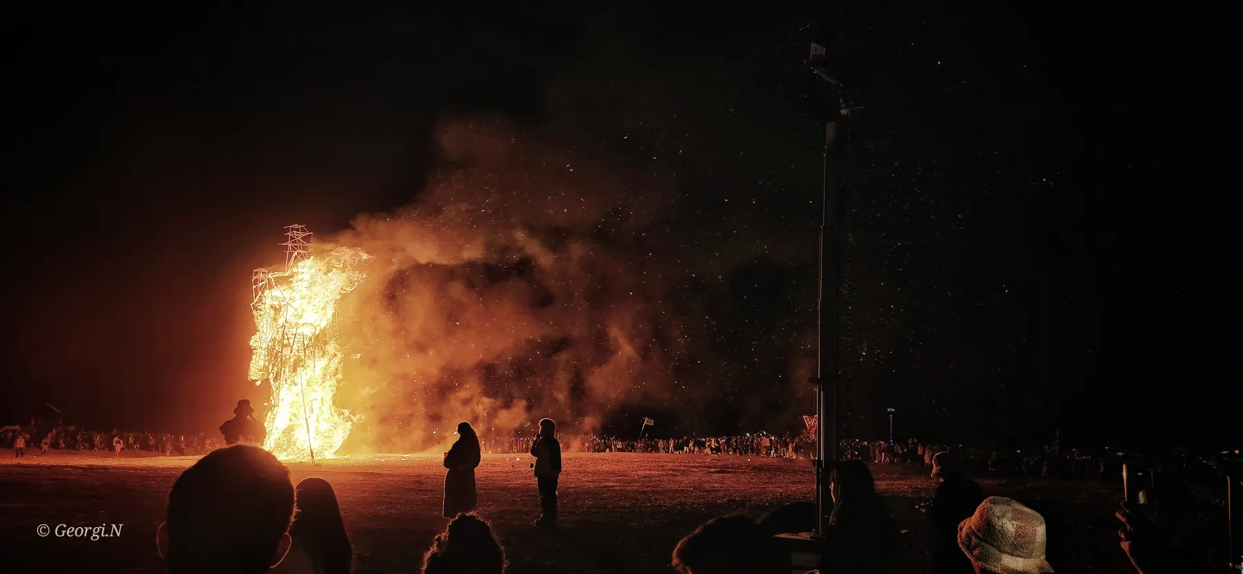 People watch a large installation burn from a distance at night.