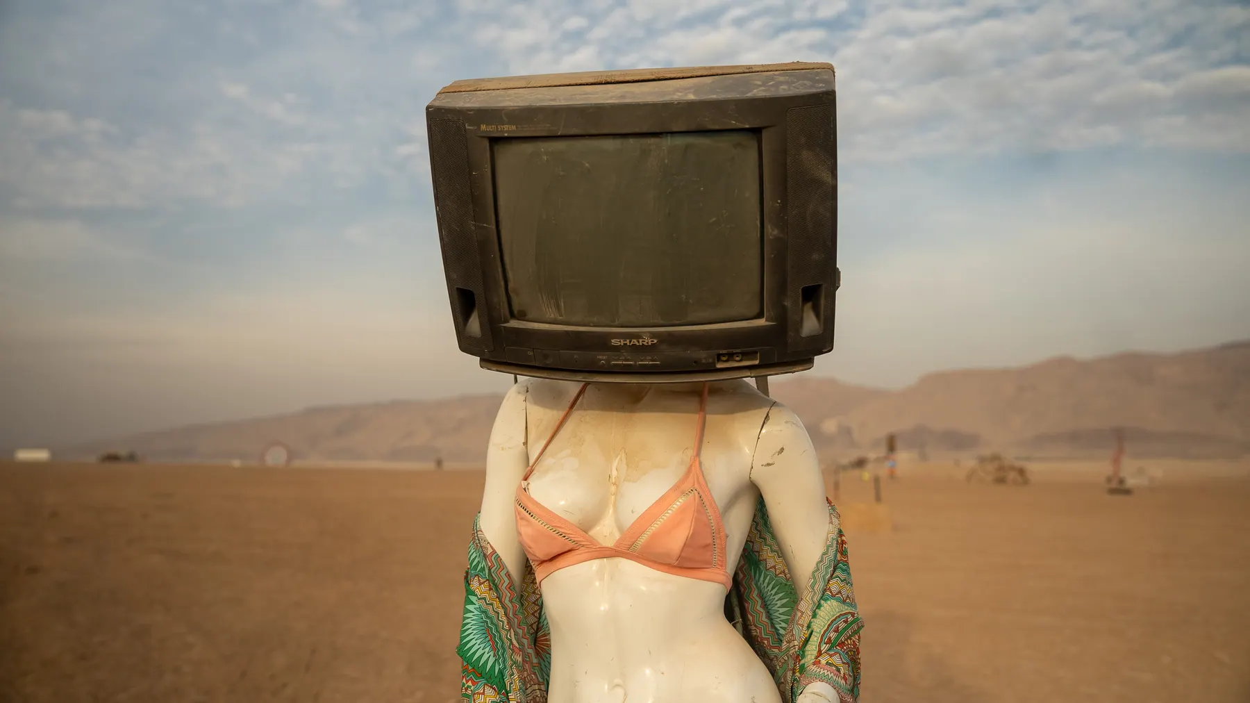 A figure with an old television set for a head stands in the desert.