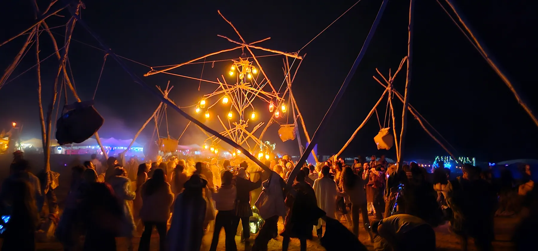 A crowd beneath branches and hanging lamps at a night gathering.