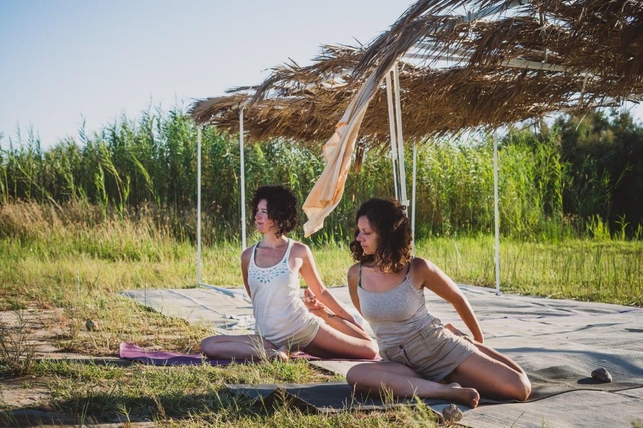 Two women sit on a mat beneath a straw shade beside the beach.