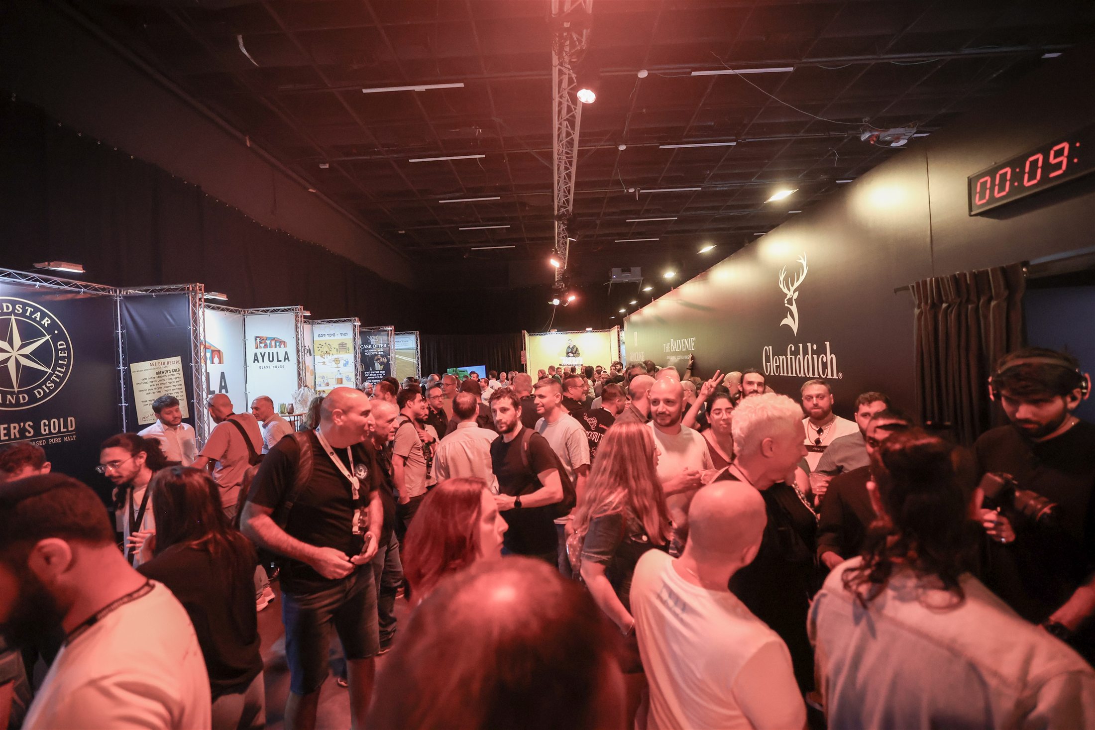 A crowd gathers around whisky stands in a dim exhibition hall.